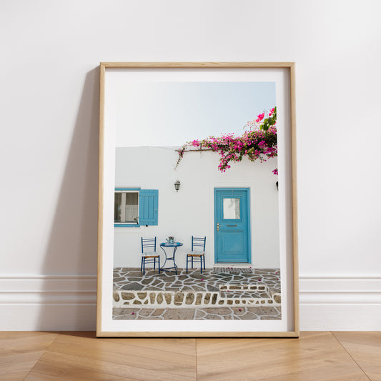 White wall with blue door and shutters, pink flowers, and outdoor furniture. ANTIPAROS, CYCLADES ISLANDS, GREECE Wall Art Prints. Adorn your walls with quaint Greek Islands scenes from Antiparos. Shop Greece Art Prints.
