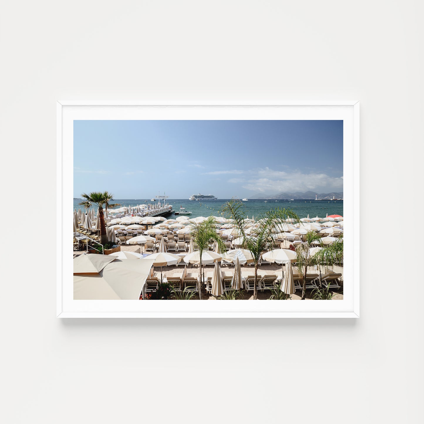 Framed photograph of a beach scene with white umbrellas and boats in the background. Bring the luxury and vibe of the celebrated Carlton Beach Club into your abode. Buy framed wall art and French Riviera Fine Art Prints. Cannes Carlton Hotel.