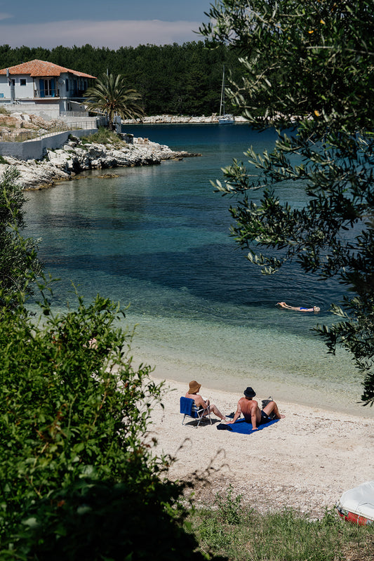 Two people sitting on a beach with a house and trees in the background. KEFALONIA, IONIAN ISLANDS, GREECE Framed Wall Art featuring the dreamy seaside holiday village of Fiscardo. Ionian Islands Fine Art Framed Prints.