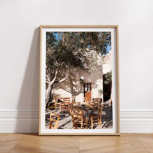 Framed photograph of an outdoor setting with tables and chairs under a tree. SHOP MONEMVASIA, GREECE | Greece Art Photography Prints, bring the warmth of Greece cafe life in to your home. Greece Cafe Wall Art.