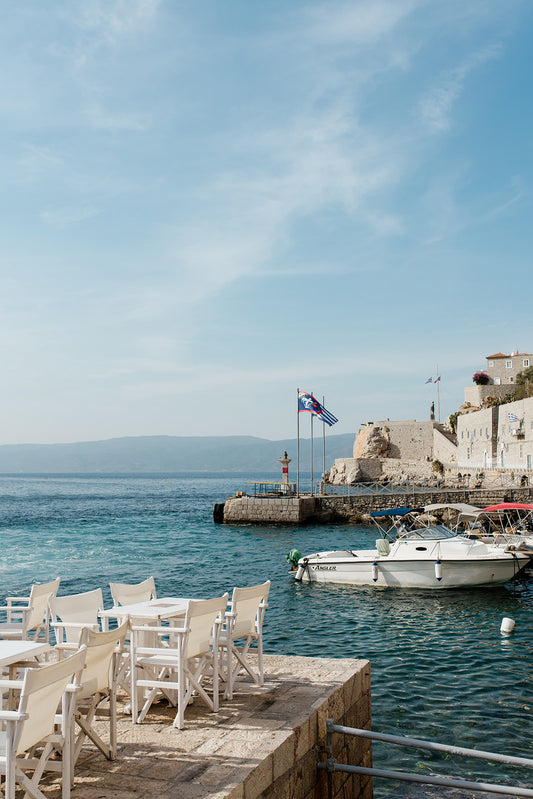 Photograph of a coastal scene with chairs on a dock and a boat in the water. HYDRA ISLAND, GREECE. SHOP Mediterranean Fine Art Prints with scenes of Hydra Island and bring the summer glow into your home. Hydra Island Wall Art.