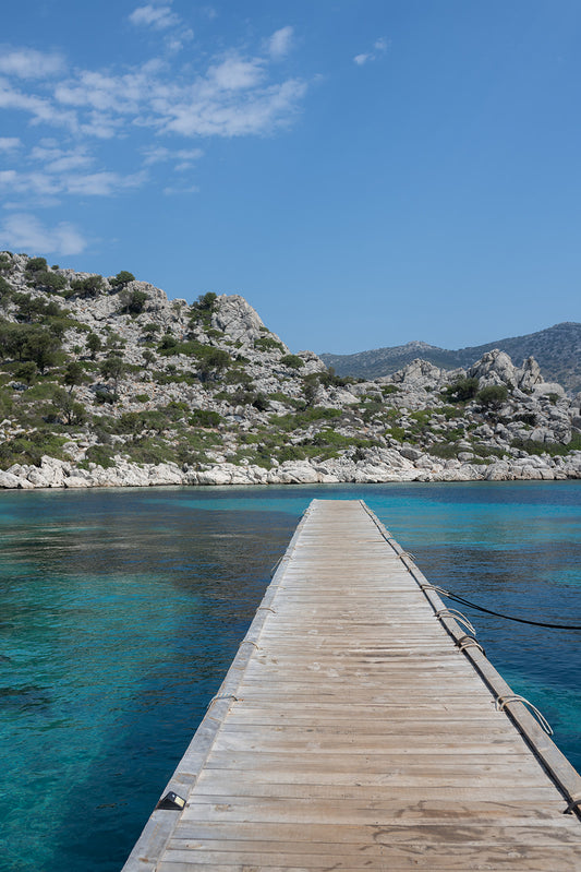 Framed photograph of a wooden dock extending into clear blue water with a mountainous background. DALYAN, TURKIYE, GREECE | Browse our range of fine art travel prints that bring the calm and space of exploration into your home. Mediterranean Wall Art.