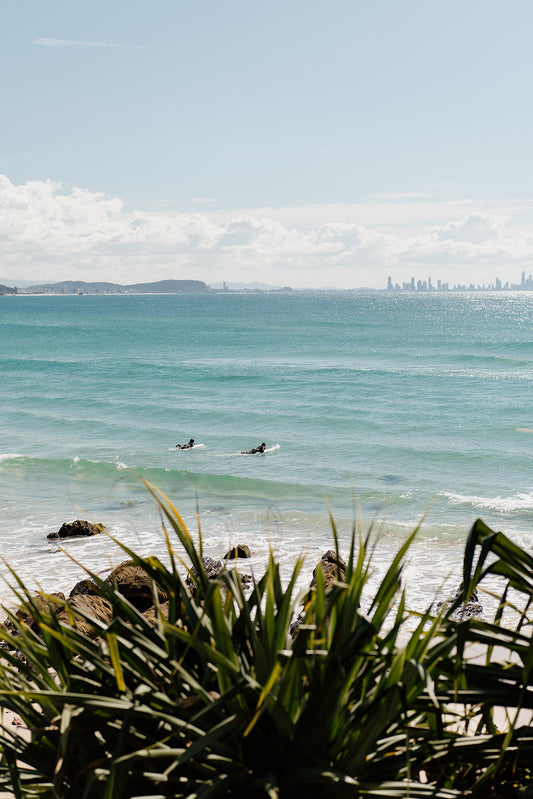 Two surfers in the ocean with a city skyline in the distance, viewed from a coastal area with greenery. Framed artwork of a beach scene with ocean and sky. KIRRA BEACH, GOLD COAST, AUSTRALIA Australia Coastal Fine Art Photography Prints by Brooke Darling Photography. Bring the endless Australian summer into your home.