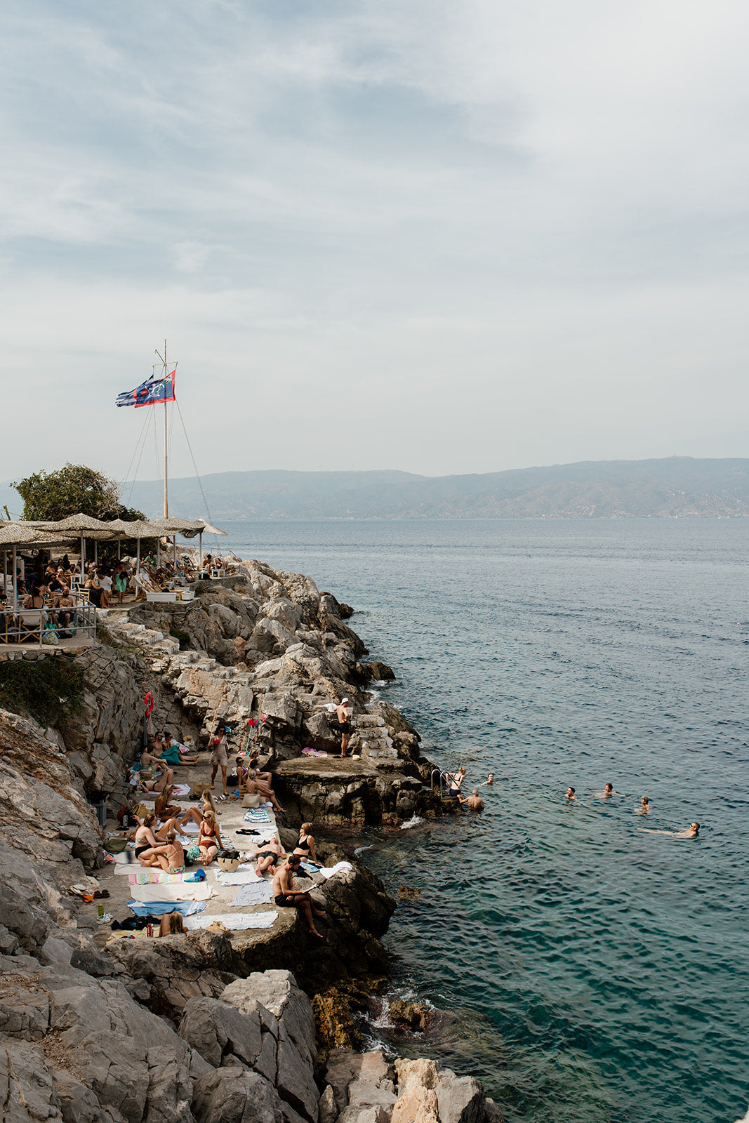 Fine Art photograph of a rocky coastline with people and a flag, set against a blue sky. HYDRA ISLAND, GREECE. SHOP our range of Mediterranean Fine Art Prints. Bring the European summer vibes in your home, year round. Mediterranean Wall Art.