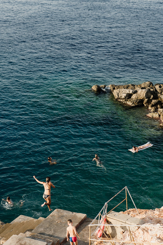 Art photograph of people swimming and diving into the ocean from a rocky shore. HYDRA ISLAND, GREECE. SHOP | European Fine Art Photography Prints. Bring the spirit of European summers into your space. Europe Summer Photography Wall Art.