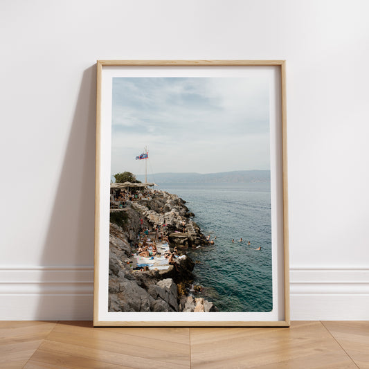 Framed photograph of a rocky coastline with people and a flag, set against a blue sky. HYDRA ISLAND, GREECE. SHOP our range of Mediterranean Fine Art Prints. Bring the European summer vibes in your home, year round. Mediterranean Wall Art.