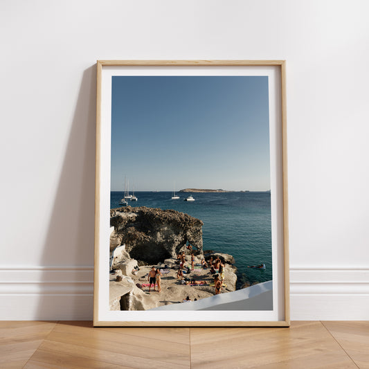 Photograph of a scenic coastal view with people on rocks and boats in the distance. Shop Greece Art Prints with Kimolos friends on a rock.