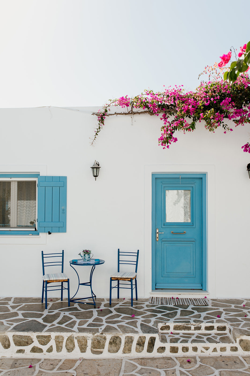 White wall with blue door and shutters, pink flowers, and outdoor furniture. ANTIPAROS, CYCLADES ISLANDS, GREECE Wall Art Prints. Adorn your walls with quaint Greek Islands scenes from Antiparos. Shop Greece Art Prints. 
