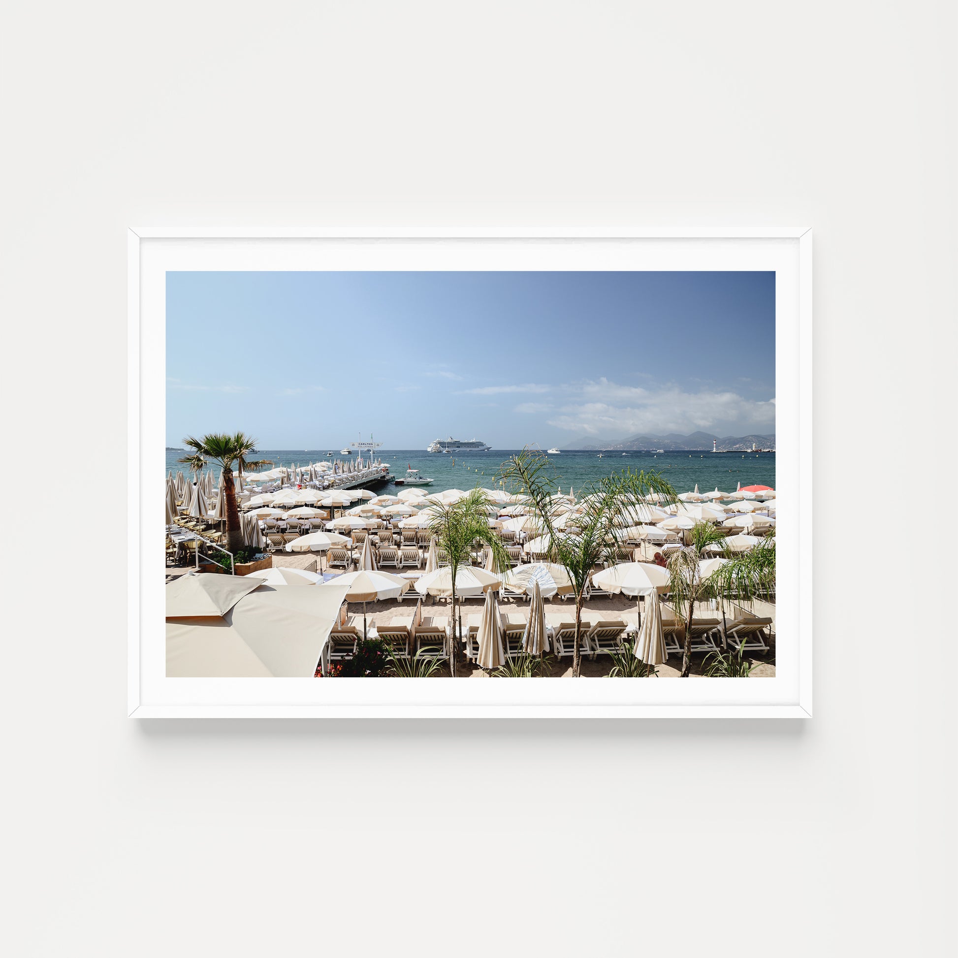 Framed photograph of a beach scene with white umbrellas and boats in the background. Bring the luxury and vibe of the celebrated Carlton Beach Club into your abode. Buy framed wall art and French Riviera Fine Art Prints. Cannes Carlton Hotel. 