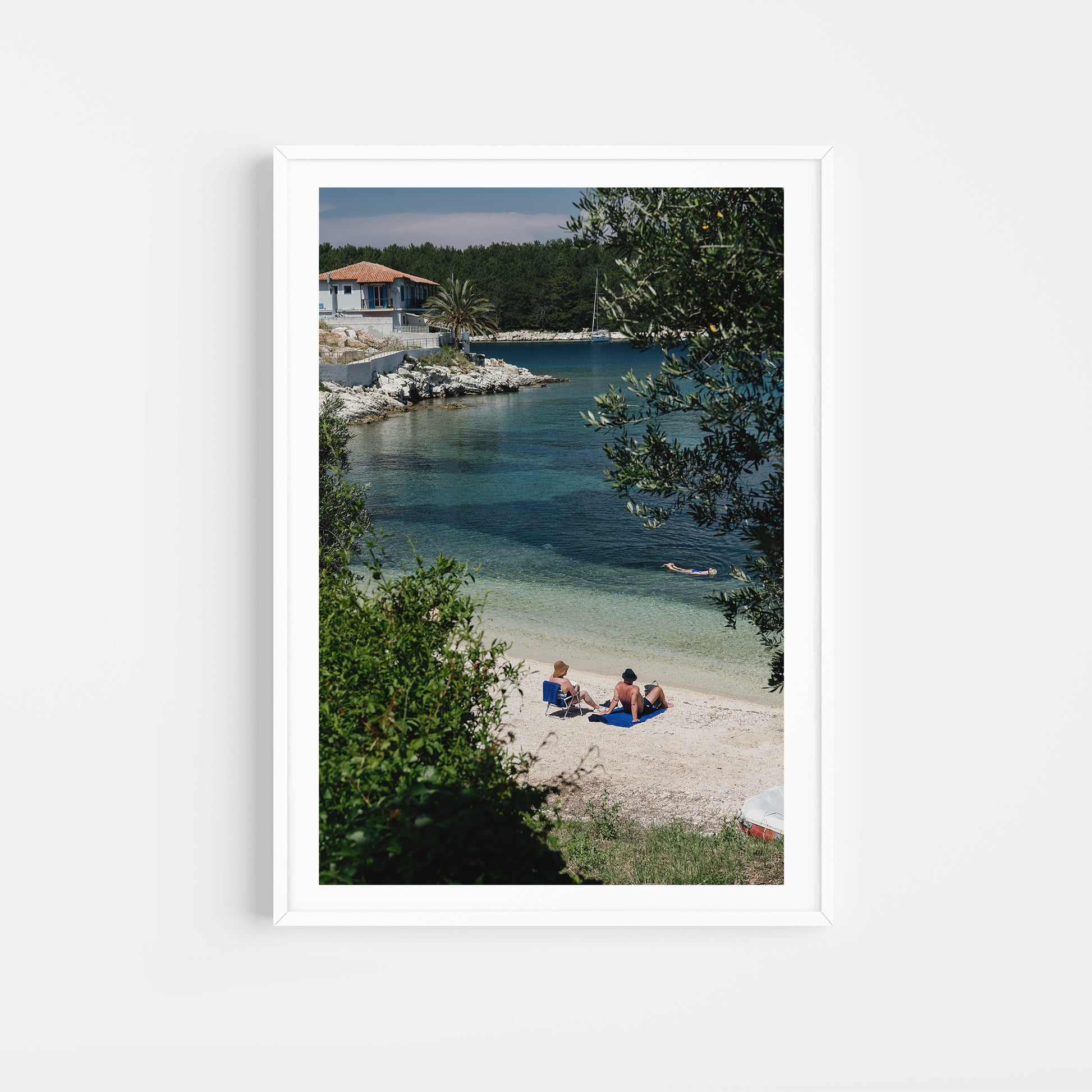 Two people sitting on a beach with a house and trees in the background. KEFALONIA, IONIAN ISLANDS, GREECE Framed Wall Art featuring the dreamy seaside holiday village of Fiscardo. Ionian Islands Fine Art Framed Prints. 