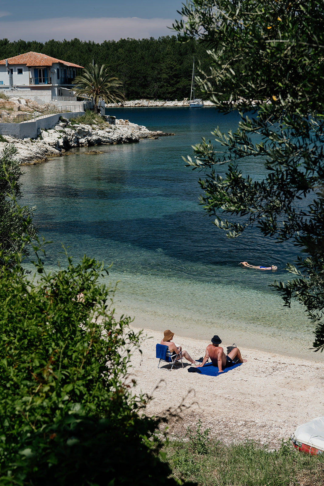 Two people sitting on a beach with a house and trees in the background. KEFALONIA, IONIAN ISLANDS, GREECE Framed Wall Art featuring the dreamy seaside holiday village of Fiscardo. Ionian Islands Fine Art Framed Prints. 