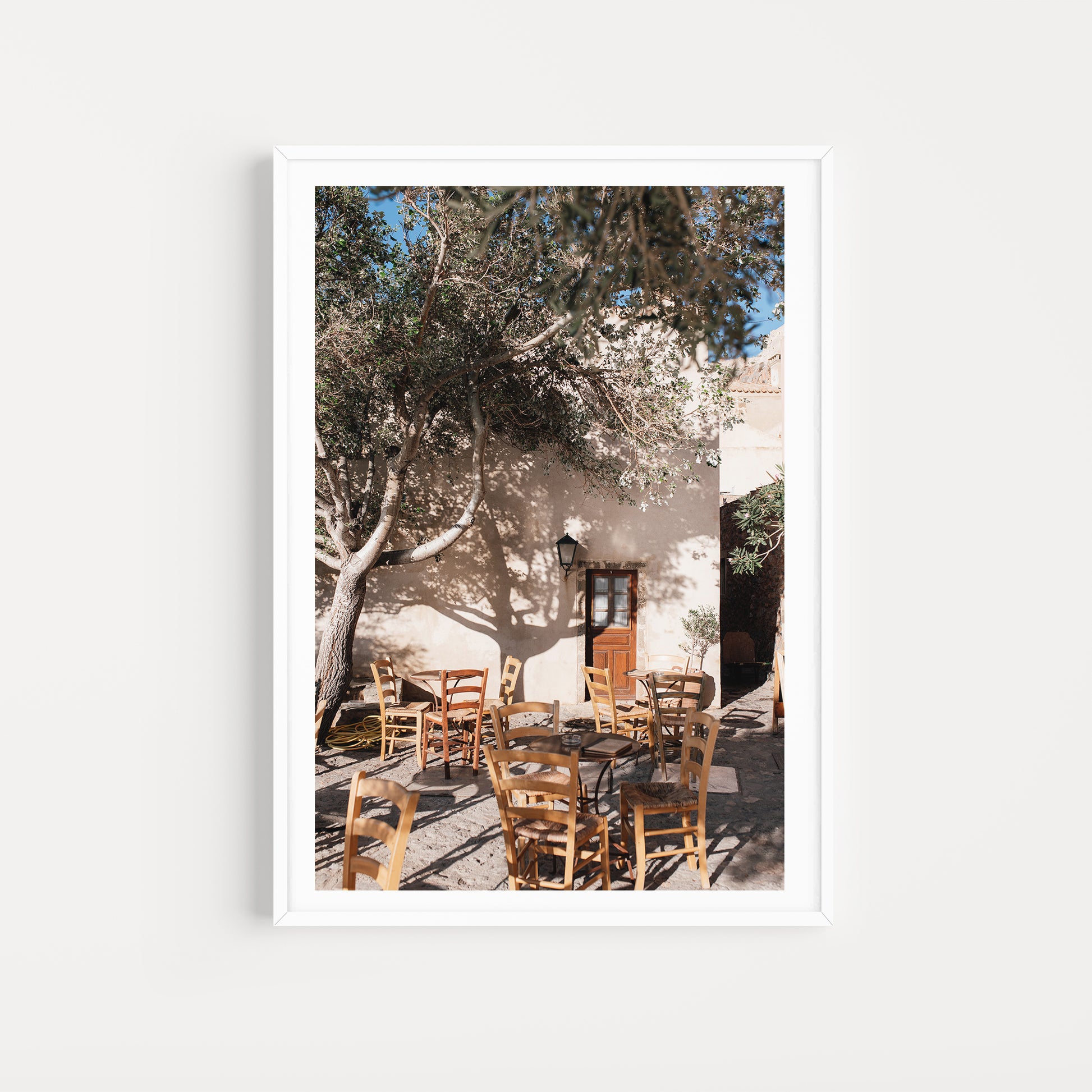 Framed photograph of an outdoor setting with tables and chairs under a tree. SHOP MONEMVASIA, GREECE | Greece Art Photography Prints, bring the warmth of Greece cafe life in to your home. Greece Cafe Wall Art. 