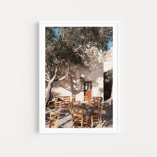 Framed photograph of an outdoor setting with tables and chairs under a tree. SHOP MONEMVASIA, GREECE | Greece Art Photography Prints, bring the warmth of Greece cafe life in to your home. Greece Cafe Wall Art. 