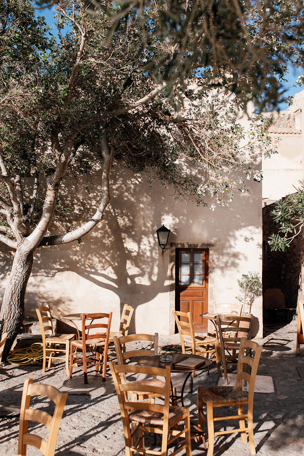 Art photograph of an outdoor setting with tables and chairs under a tree. SHOP MONEMVASIA, GREECE | Greece Art Photography Prints, bring the warmth of Greece cafe life in to your home. Greece Cafe Wall Art. 