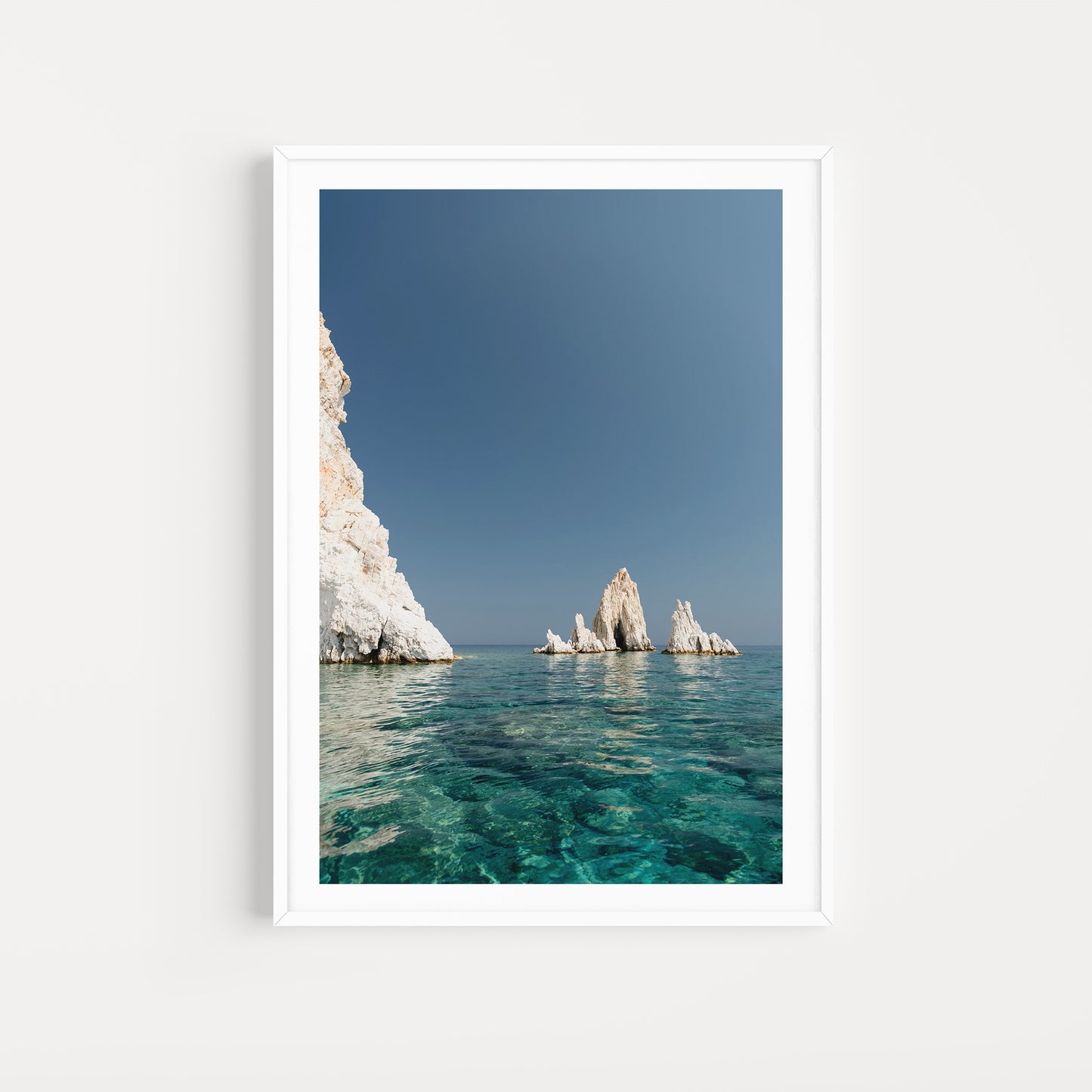 Greek Islands art print schowcasing vibrant blue portrait image of rocks at and ocean at Polyaigos Island near Milos. White frame mock up of Greece fine art photographic print.