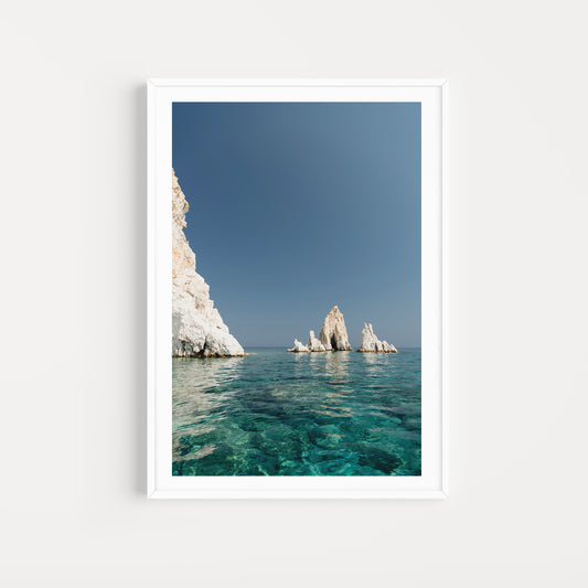 Greek Islands art print schowcasing vibrant blue portrait image of rocks at and ocean at Polyaigos Island near Milos. White frame mock up of Greece fine art photographic print.