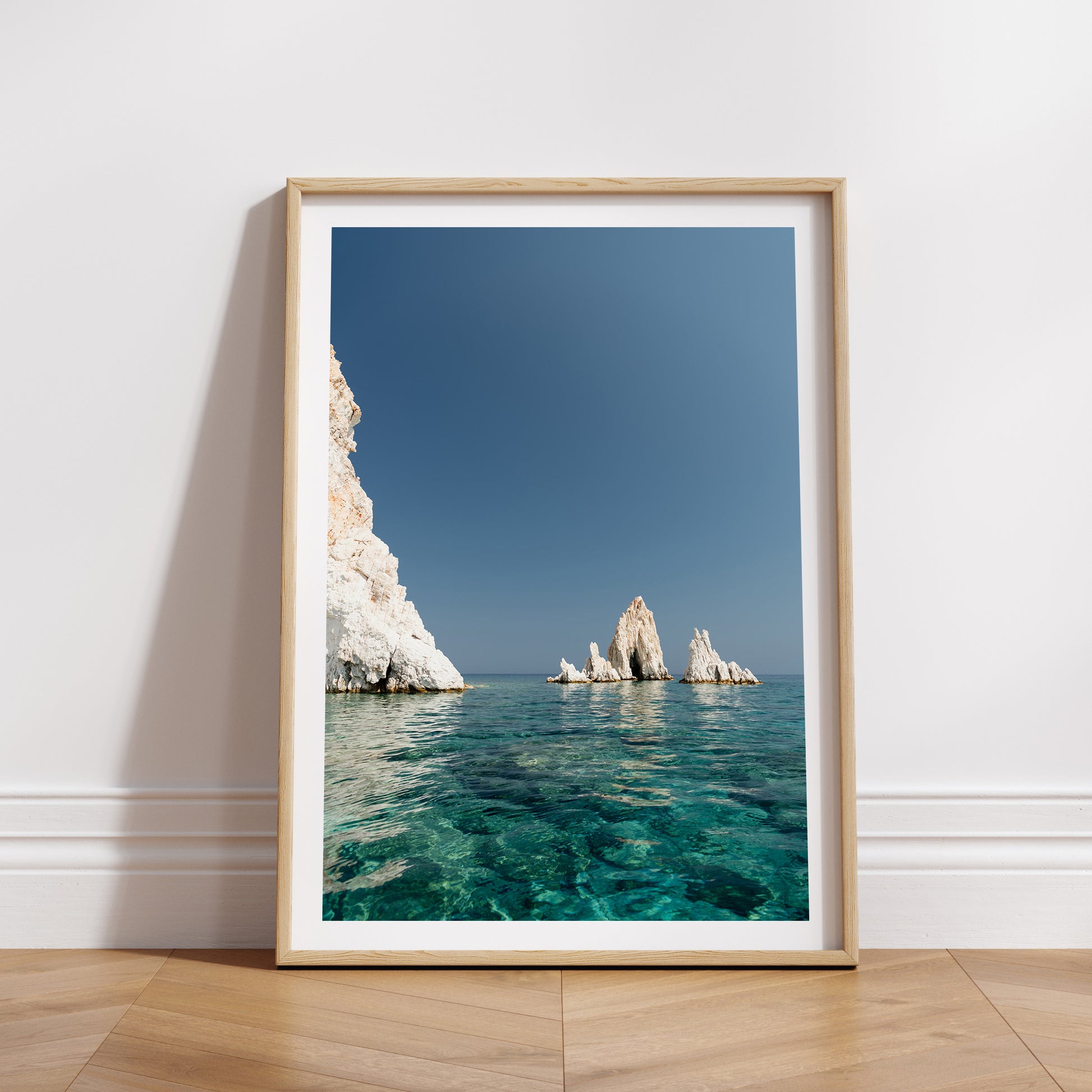 Greek Islands art print schowcasing vibrant blue portrait image of rocks at and ocean at Polyaigos Island near Milos. Wood frame mock up of Greece fine art photographic print.