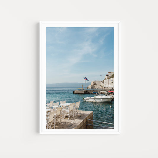 Framed photograph of a coastal scene with chairs on a dock and a boat in the water. HYDRA ISLAND, GREECE. SHOP Mediterranean Fine Art Prints with scenes of Hydra Island and bring the summer glow into your home. Hydra Island Wall Art.