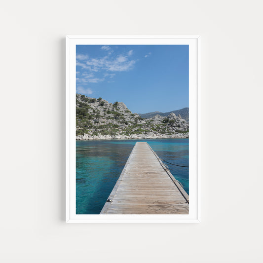 Framed photograph of a wooden dock extending into clear blue water with a mountainous background. DALYAN, TURKIYE, GREECE | Browse our range of fine art travel prints that bring the calm and space of exploration into your home. Mediterranean Wall Art. 
