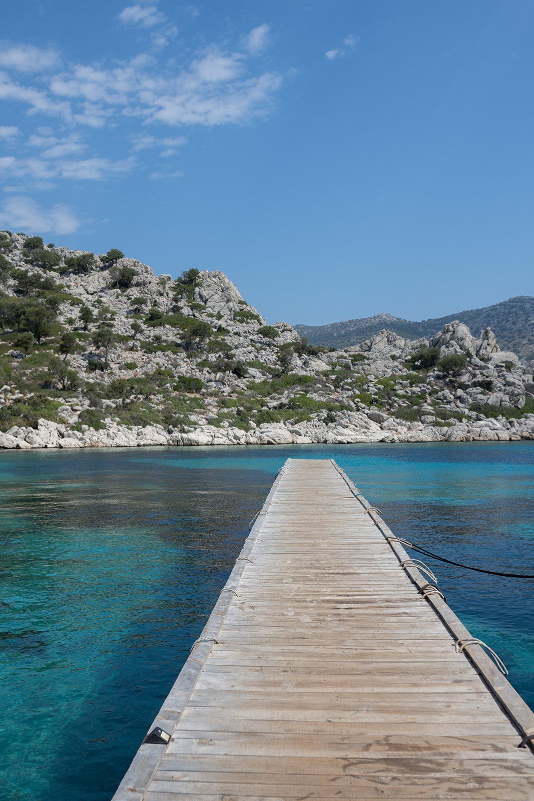 Framed photograph of a wooden dock extending into clear blue water with a mountainous background. DALYAN, TURKIYE, GREECE | Browse our range of fine art travel prints that bring the calm and space of exploration into your home. Mediterranean Wall Art. 