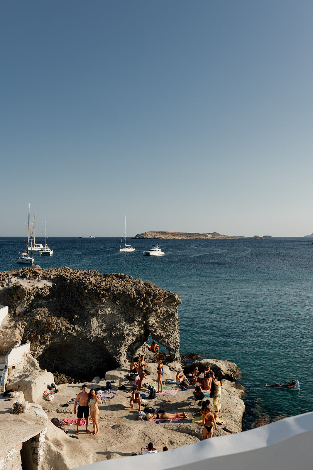 Photograph of a scenic coastal view with people on rocks and boats in the distance. Shop Greece Art Prints with Kimolos friends on a rock. 