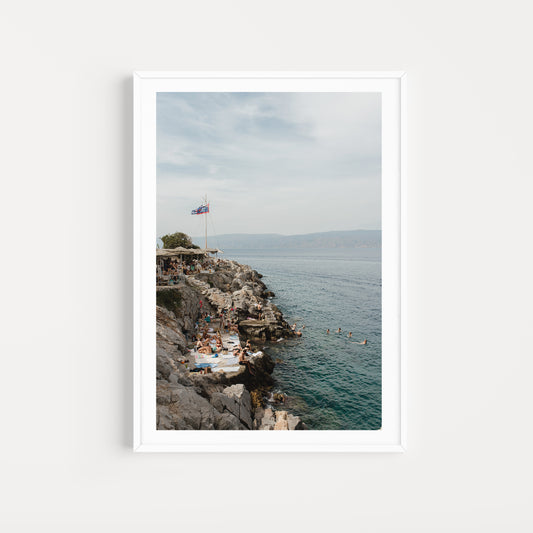 Framed photograph of a rocky coastline with people and a flag, set against a blue sky. HYDRA ISLAND, GREECE. SHOP our range of Mediterranean Fine Art Prints. Bring the European summer vibes in your home, year round. Mediterranean Wall Art. 